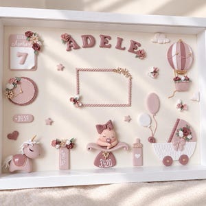 Könnte beinhalten: Ein weißer Schattenfugenrahmen mit einer 3D-Darstellung für ein Baby namens ADELE. Er enthält einen Kalender mit dem Datum 7. April 2022, eine Uhr, ein Einhorn, einen Heißluftballon, einen Kinderwagen, ein Fläschchen und eine schlafende Babyfigur.