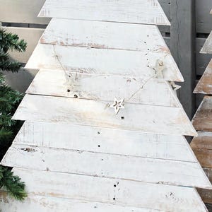 May include: A white painted wooden Christmas tree decoration with a distressed finish, featuring a galvanized metal star topper and a string of small star ornaments draped across the front, supported by a brown wooden base stand.