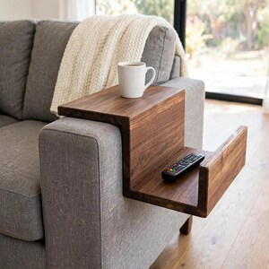 Könnte beinhalten: Ein dunkelbrauner Sofa-Armlehnentisch mit abgestuftem Design, der eine flache Oberseite mit einer weißen Keramiktasse und eine untere Ablage mit einer schwarzen Fernbedienung besitzt und fest auf der Armlehne eines grauen Sofas aufliegt.