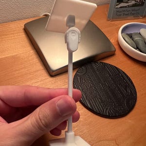 May include: A white plastic stand featuring a square base, a thin vertical rod, and a small square platform at the top. The platform is connected to the rod by a joint mechanism that allows for adjustment. The entire structure is made of white material.