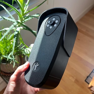 May include: A black, textured, vertical rectangular device with a rounded top and a protective hood. The front features a central camera lens surrounded by five small circular sensors and a small, light-colored Y-shaped logo at the bottom.