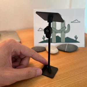 May include: A black adjustable stand featuring a square base, a thin vertical pole, and a flat rectangular top platform. The stand includes a central hinge mechanism with a small circular logo detail on the front of the joint.