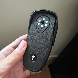 May include: A black, textured, rectangular electronic device with rounded corners. The top section features a circular array of five white dots surrounding a central hole. The bottom section displays a small white logo resembling a stylized plant sprout.