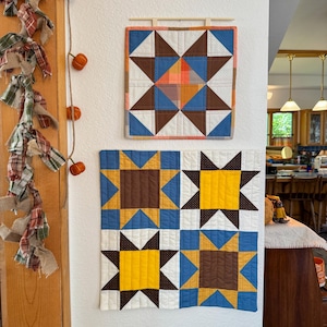 May include: Two square quilted wall hangings featuring geometric star patterns in shades of brown, blue, yellow, and white. The top quilt has a single large star, while the bottom quilt is divided into four quadrants, each containing a smaller star.
