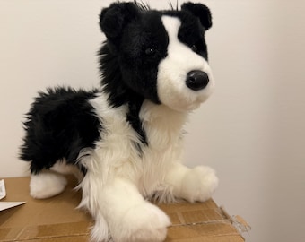 Puede incluir: Un peluche con forma de perro border collie. El juguete presenta un pelaje largo y suave de color blanco y negro, una mancha blanca en la cara, ojos oscuros y una nariz negra. Está representado en una posición sentada con las patas.