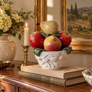 May include: A white ceramic bowl with embossed floral details holds a collection of realistic ceramic fruit, including a speckled yellow pear, a yellow and red peach, and two bright red apples, all accented with green ceramic leaves.