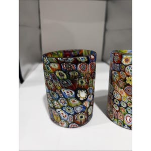 May include: Two cylindrical glass tumblers featuring a dense, multicolored millefiori pattern. The surface is covered in small, intricate floral-like glass canes in shades of red, blue, green, yellow, and white, creating a vibrant mosaic effect.