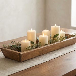 May include: A long, rectangular wooden dough bowl filled with five lit cream-colored pillar candles of varying heights, accented with sprigs of green eucalyptus leaves tucked between the candles inside the wooden container.