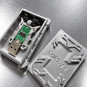 Könnte beinhalten: Ein silberfarbenes, 3D-gedrucktes rechteckiges Gehäuse mit passendem Deckel, der den Text CORE38 trägt. Im Inneren befindet sich ein USB-Stick mit grüner Leiterplatte und eine schwarze Perlenketten-Halskette im Fach.