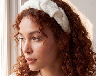 Puede incluir: Una diadema de punto gruesa en color blanco roto que presenta un prominente patrón de trenzado. El accesorio está confeccionado con un hilo suave y texturizado, y se coloca sobre la cabeza mostrando un diseño tejido voluminoso con puntos visibles.