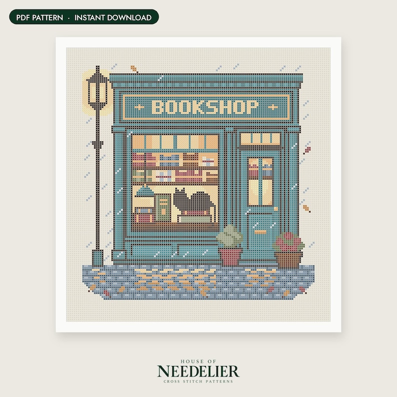 May include: A cross-stitch pattern featuring a teal bookstore storefront with the word BOOKSHOP above the window. The design includes a black cat in the window, a tall street lamp, potted plants, and a cobblestone sidewalk with scattered leaves.