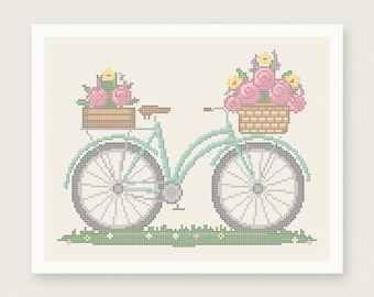 May include: A cross stitch pattern featuring a light blue bicycle with a brown wooden crate on the back and a woven basket on the front, both filled with pink and yellow flowers. The bicycle sits on a green grassy patch with small flowers.