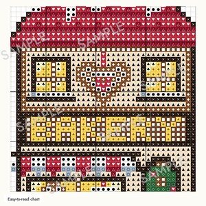 May include: A cross-stitch pattern chart featuring a bakery building design. The grid displays a red roof, two windows, a central heart, the word BAKERY in yellow, and a striped awning over a storefront with a green door, all using symbols.