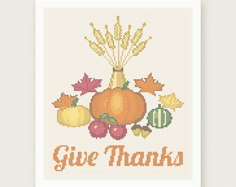 May include: A cross stitch pattern featuring a central orange pumpkin, a yellow pumpkin, a green striped gourd, red apples, acorns, and autumn leaves. Above the produce is a bundle of wheat stalks. The text "Give Thanks" is in orange below.