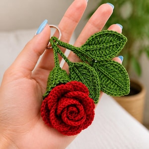 May include: A crochet keyring featuring a vibrant red rose flower attached to a silver metal ring. The rose is accompanied by three detailed green crocheted leaves and a green stem, all crafted with intricate yarn stitching.