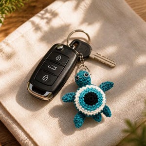 May include: A black car key fob attached to a metal keyring with a small, handmade crochet turtle keychain. The turtle features a teal body and a circular shell design with concentric rings of white, light blue, and a black center.