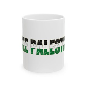 May include: A white ceramic mug featuring the text FREE PALESTINE printed in a distressed, weathered font. The letters are filled with horizontal stripes of black, white, and green, creating a textured, worn appearance against the plain white surface.