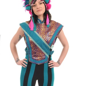 May include: A woman wearing a teal and black striped pants with a teal, gold, and purple patterned vest. She is wearing a pink and teal feathered headdress with pink pom-poms.