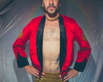 Könnte beinhalten: Eine Person trägt eine rote Satinjacke mit schwarzem Samtbesatz und goldenen Akzenten. Die Jacke ist offen und zeigt eine nackte Brust mit einem Tattoo. Die Person trägt eine goldene Hose.