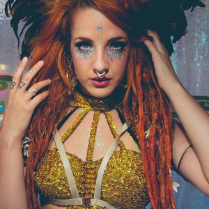 K&ouml;nnte beinhalten: Eine Frau mit roten Haaren und Dreadlocks tr&auml;gt ein goldenes Paillettenoberteil mit einem wei&szlig;en Gurtzeug. Sie tr&auml;gt ein Einhorn-Stirnband mit Federn und hat Glitzer-Make-up im Gesicht.