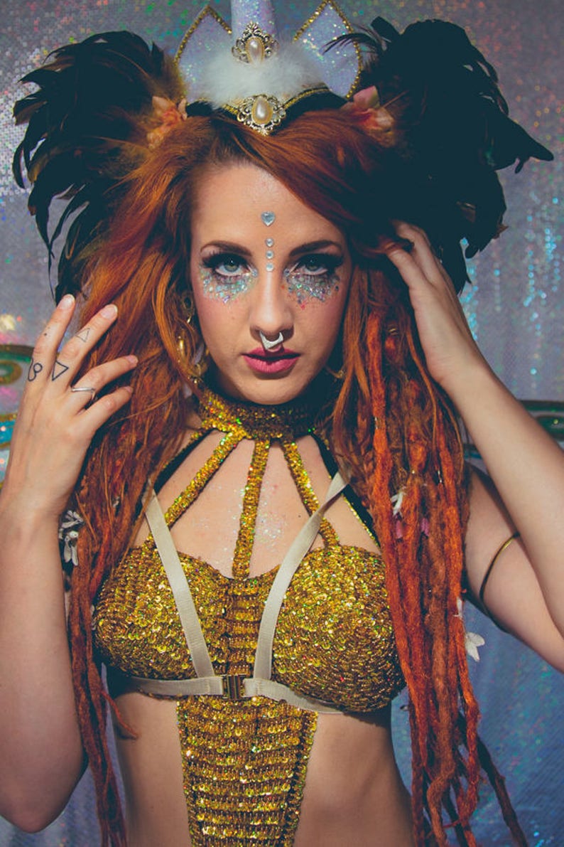 K&ouml;nnte beinhalten: Eine Frau mit roten Haaren und Dreadlocks tr&auml;gt ein goldenes Paillettenoberteil mit einem wei&szlig;en Gurtzeug. Sie tr&auml;gt ein Einhorn-Stirnband mit Federn und hat Glitzer-Make-up im Gesicht.