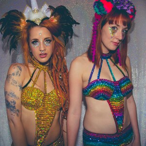 K&ouml;nnte beinhalten: Zwei Frauen tragen farbenfrohe, paillettenbesetzte Oberteile und Stirnb&auml;nder. Eine Frau tr&auml;gt ein goldenes Oberteil mit einem Einhorn-Stirnband, die andere ein Regenbogen-Oberteil mit einem Blumen-Stirnband.