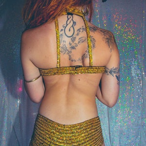 K&ouml;nnte beinhalten: Eine Frau tr&auml;gt einen goldenen Pailletten-BH und passende Shorts. Sie hat ein Tattoo auf dem R&uuml;cken mit einem Knoten und Bl&auml;ttern.