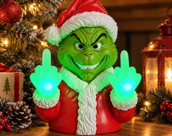 Könnte beinhalten: Eine grüne Charakterfigur in einem roten Weihnachtsmannkostüm mit weißem Pelzbesatz und passender Mütze. Die Figur hat einen schelmischen Gesichtsausdruck und hält beide Hände mit leuchtend grünen, ausgestreckten Mittelfingern hoch.
