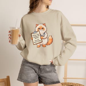 May include: A beige crewneck sweatshirt featuring a graphic of a fluffy orange and white cat wearing a stethoscope and holding a clipboard that reads, "Please stop copying and pasting. Sincerely, Medical Coders." The sweatshirt has long sleeves.