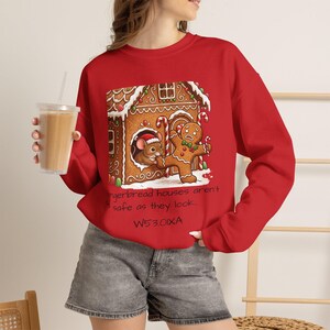 May include: A red long-sleeve sweatshirt featuring a graphic of a gingerbread house, a mouse, and a gingerbread person. The text reads: Gingerbread houses aren't safe as they look... W53.0IXA. The design includes white icing details and candy canes.