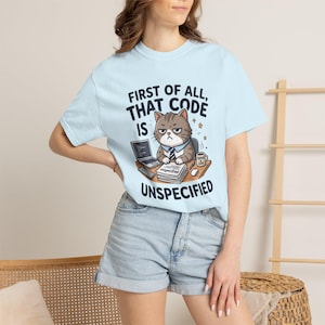 Può includere: Una t-shirt azzurra con la grafica di un gatto imbronciato seduto a una scrivania con un portatile, una pila di fogli con scritto unspecified e una tazza di caffè. Il testo recita FIRST OF ALL, THAT CODE IS UNSPECIFIED in lettere blu scuro.