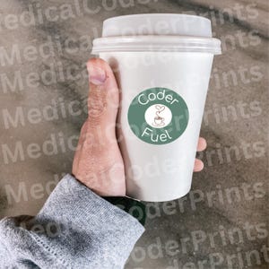 Pode incluir: Um copo de café descartável branco com tampa de plástico branca. Um autocolante circular verde-sálvia está centrado no copo, apresentando o texto Coder Fuel em letras brancas e uma pequena ilustração de linha de uma chávena de café.