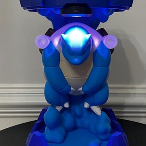 May include: A blue and white creature figurine standing on a blue base, featuring a glowing light on its head and a dark blue structure overhead. The figure has a rounded body, white claws, and a smooth matte finish, illuminated by internal LEDs.