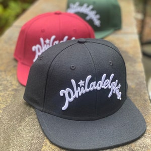 May include: Three baseball caps in black, burgundy, and green, each embroidered with the word "Philadelphia" in white script. The caps are displayed on a stone surface, with the black cap in the foreground.