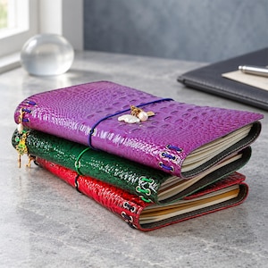 May include: A stack of three rectangular journals with textured, glossy covers in purple, green, and red. Each journal features a crocodile-skin pattern, metal eyelets with colored elastic cords, and small decorative charms attached to the side.