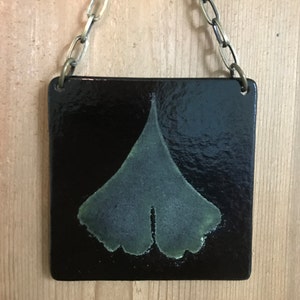 May include: A black ceramic tile with a green ginkgo leaf design hangs from a brass chain.