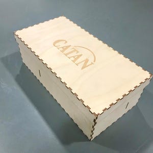 May include: A light-colored wooden box with the word "CATAN" etched on the lid. The box has a rectangular shape with a notched edge design. The box is likely for storing a board game.
