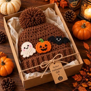 May include: A brown crocheted beanie with a large pom-pom, featuring appliqués of a white ghost, an orange jack-o'-lantern, and a black bat. The beanie is inside a cardboard gift box with a tag that reads Perfect Halloween Gift with a pumpkin icon.