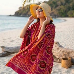 Op de afbeelding: Een strooien zonnehoed met een brede rand en een gele onderkant, gecombineerd met een levendige magenta en oranje sjaal met geometrische ikat-patronen en een Griekse rand langs de zoom, gedragen over een bijpassend magenta en oranje kledingstuk.