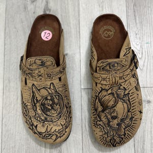 May include: A pair of tan slip-on clogs with brown footbeds and adjustable straps. The left shoe features a cat in a space helmet, while the right shoe displays a skull and crossbones. Both shoes have intricate black line art designs.