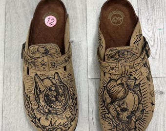 May include: A pair of tan slip-on clogs with brown footbeds and adjustable straps. The left shoe features a cat in a space helmet, while the right shoe displays a skull and crossbones. Both shoes have intricate black line art designs.
