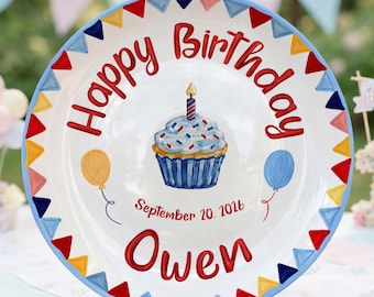 May include: A white ceramic plate featuring a hand-painted cupcake with a single candle in the center. The text reads Happy Birthday and Owen, with the date September 20, 2026. A colorful triangular bunting border surrounds the edge of the plate.