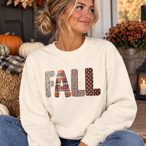 May include: A cream-coloured crewneck sweatshirt featuring the word FALL printed across the chest. Each letter has a distinct pattern: the F is floral, the A is plaid, the L is gingham, and the second L is brown with white polka dots.