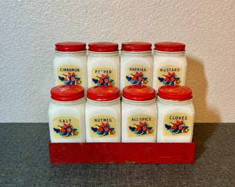 Puede incluir: Un juego de ocho frascos de especias de vidrio lechoso blanco con tapas de metal rojo, dispuestos en dos filas de cuatro en un estante de metal rojo. Cada frasco tiene una etiqueta con el nombre de la especia y una ilustración de frutas coloridas: Canela, Pimienta, Pimentón, Mostaza, Sal, Nuez moscada, Pimienta de Jamaica y Clavos.