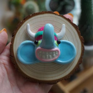 May include: A circular wood slice ornament featuring a sculpted, glossy blue elephant head with white horns, a pink mouth, and black eyes. The elephant has a prominent trunk and wide ears, mounted centrally on the natural light-colored wood grain surface.