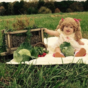 May include: A baby wearing a yarn wig with pink bows, sitting on a blanket in a field. A wooden cart, cabbages, and radishes are nearby. The baby is wearing a light-colored dress and smiling.