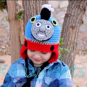 May include: A child wearing a blue, red, and gray crocheted hat with a Thomas the Tank Engine design. The hat has a black brim and a white puff on top. The child is wearing a blue plaid shirt.
