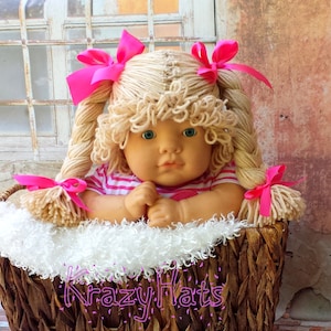 May include: A doll with blonde yarn hair styled in braids and curls, adorned with pink bows. The doll wears a pink and white striped shirt and sits in a woven basket lined with white fluff. The basket has the text "Krazy Hats".