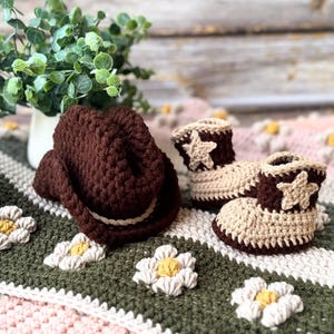 May include: A brown crochet cowboy hat and matching baby booties with star accents. The booties are tan and brown, and the hat has a white band. The items are on a floral blanket with green, white, and pink colors.