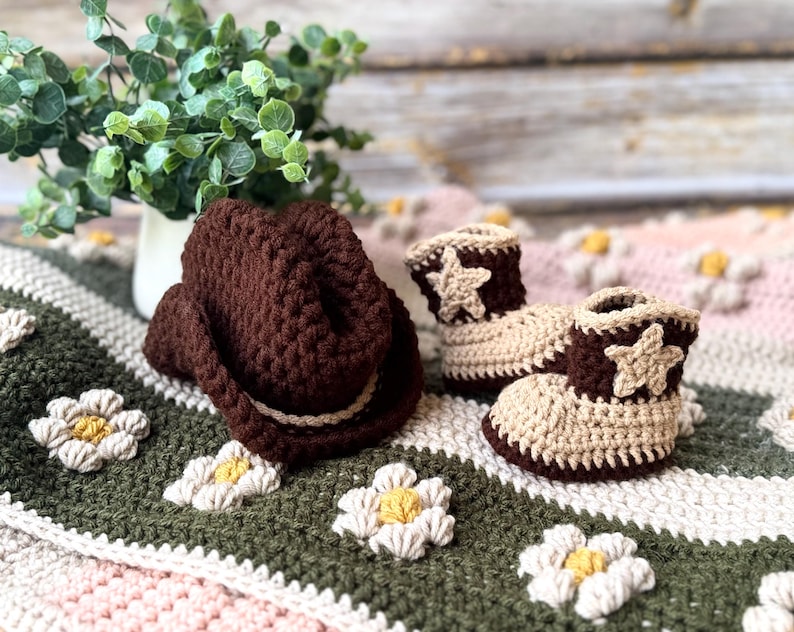May include: A brown crochet cowboy hat and matching baby booties with star accents. The booties are tan and brown, and the hat has a white band. The items are on a floral blanket with green, white, and pink colors.